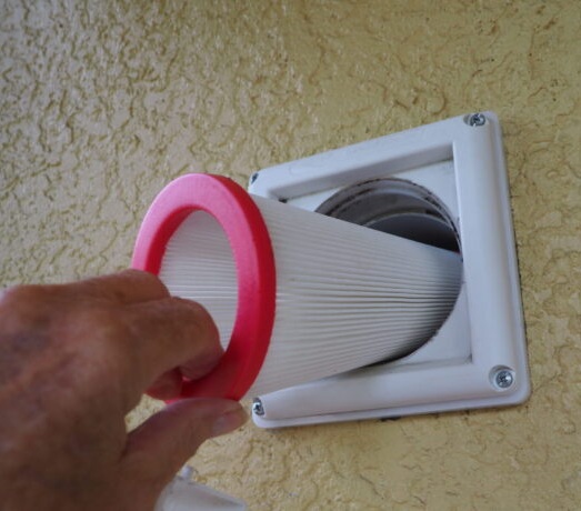 Hand inserting a rolled-up paper through a wall vent with a pink ring.
